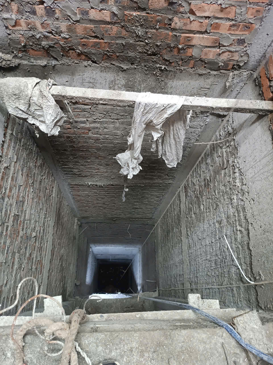 Hố thang máy bê tông đang thi công với tường gạch thô và dây cáp xây dựng